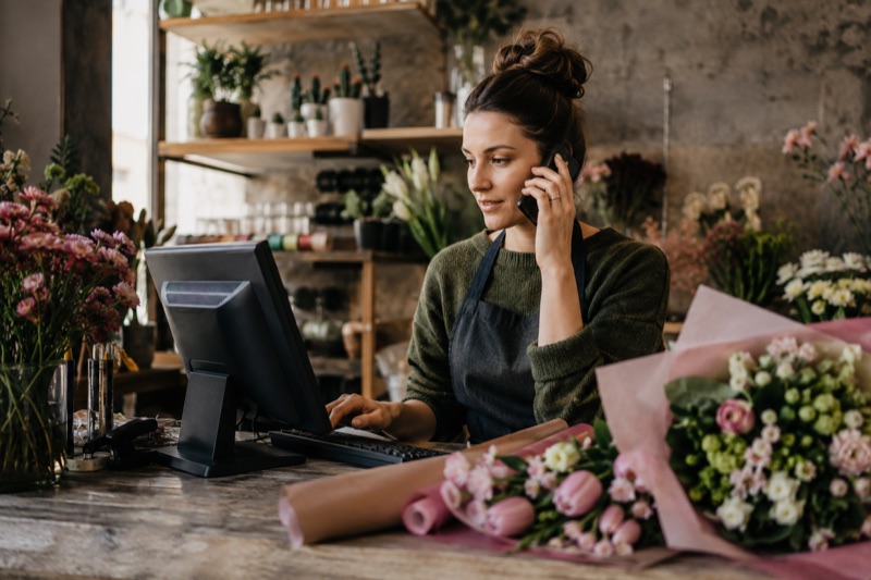 Floristă la tejgheaua florăriei, cu telefonul la ureche, notând o comandă direct în calculator, cu buchete în lucru pe masa de alături