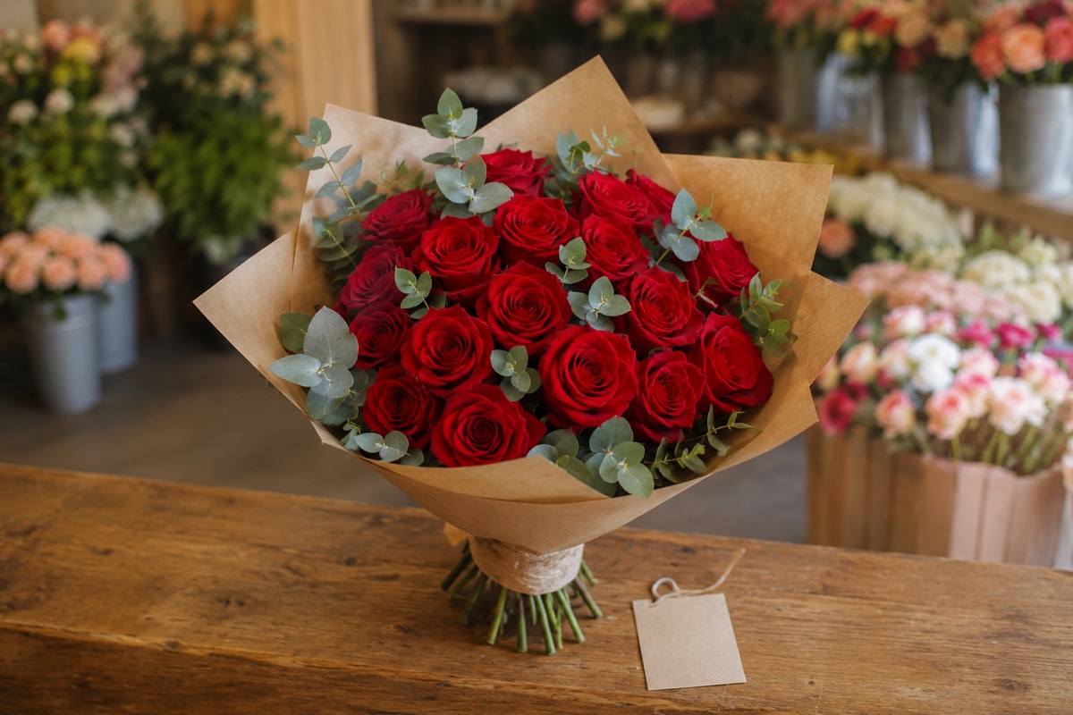 Buchet de 15 trandafiri roșii cu eucalipt, ambalat în hârtie kraft, pe tejgheaua unei florării, lângă o etichetă de preț goală